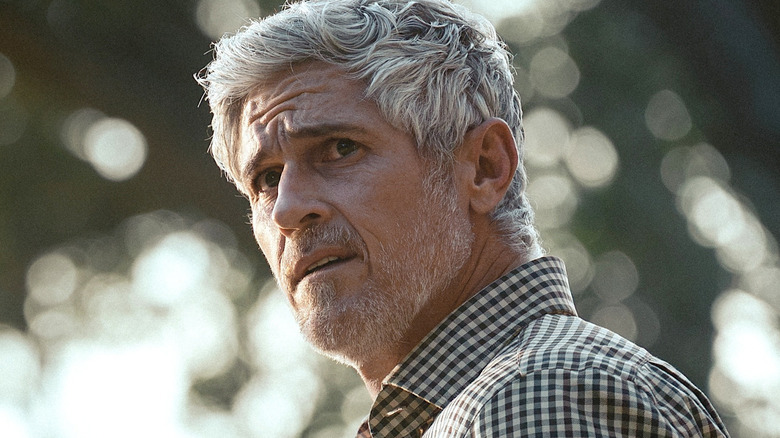 Gray-haired man with stubble in a checkered shirt looks concerned outdoors against a blurred background.