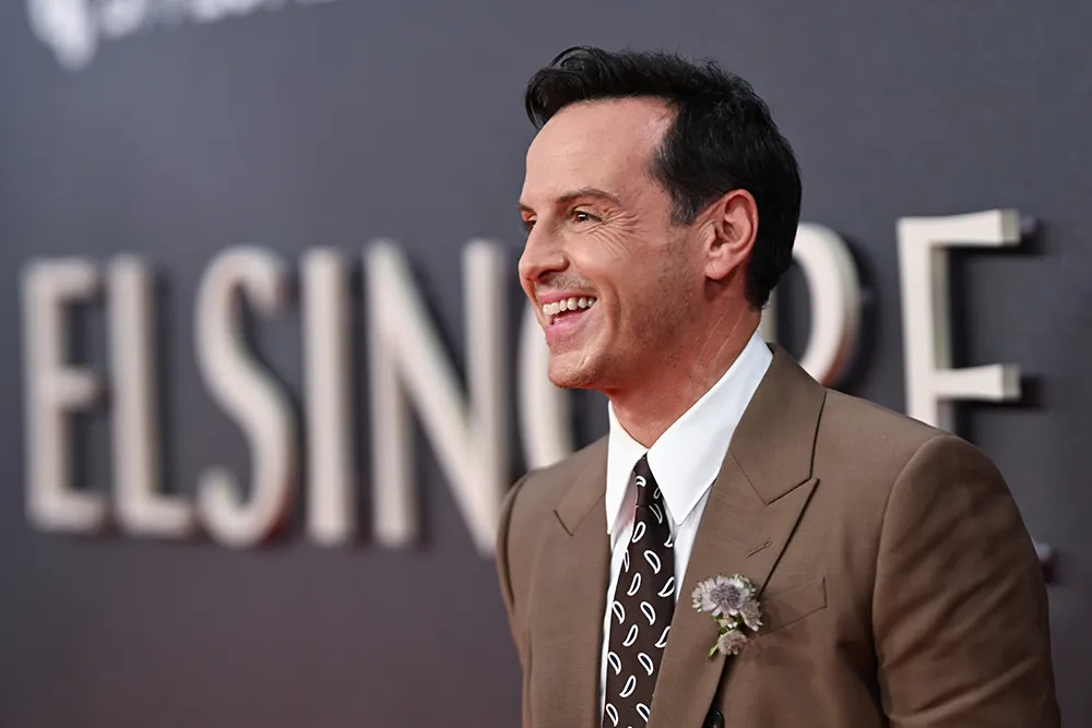A smiling man in a brown suit and patterned tie stands before a blurred ELSINORE backdrop.