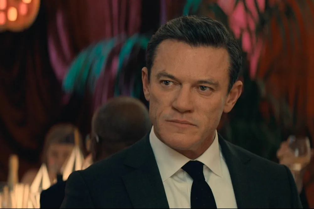 A man in a dark suit and tie looks sideways in a dimly lit, crowded room.