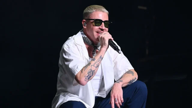 A tattooed performer in sunglasses crouches onstage, holding a microphone against a dark background.