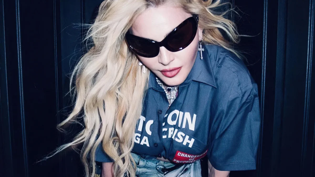 A blonde woman in oversized sunglasses leans forward against a dark paneled background.