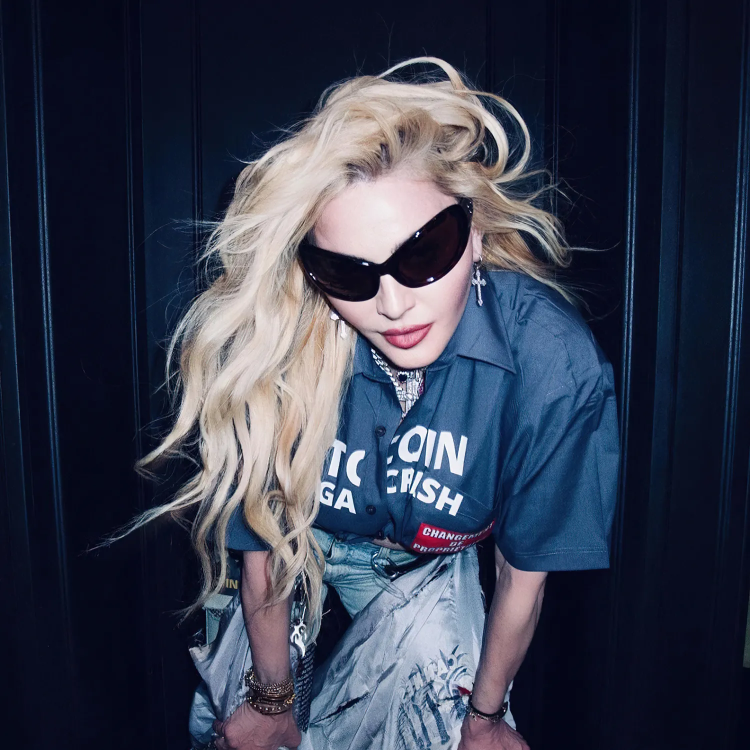 A blonde woman in oversized sunglasses leans forward against a dark paneled background.
