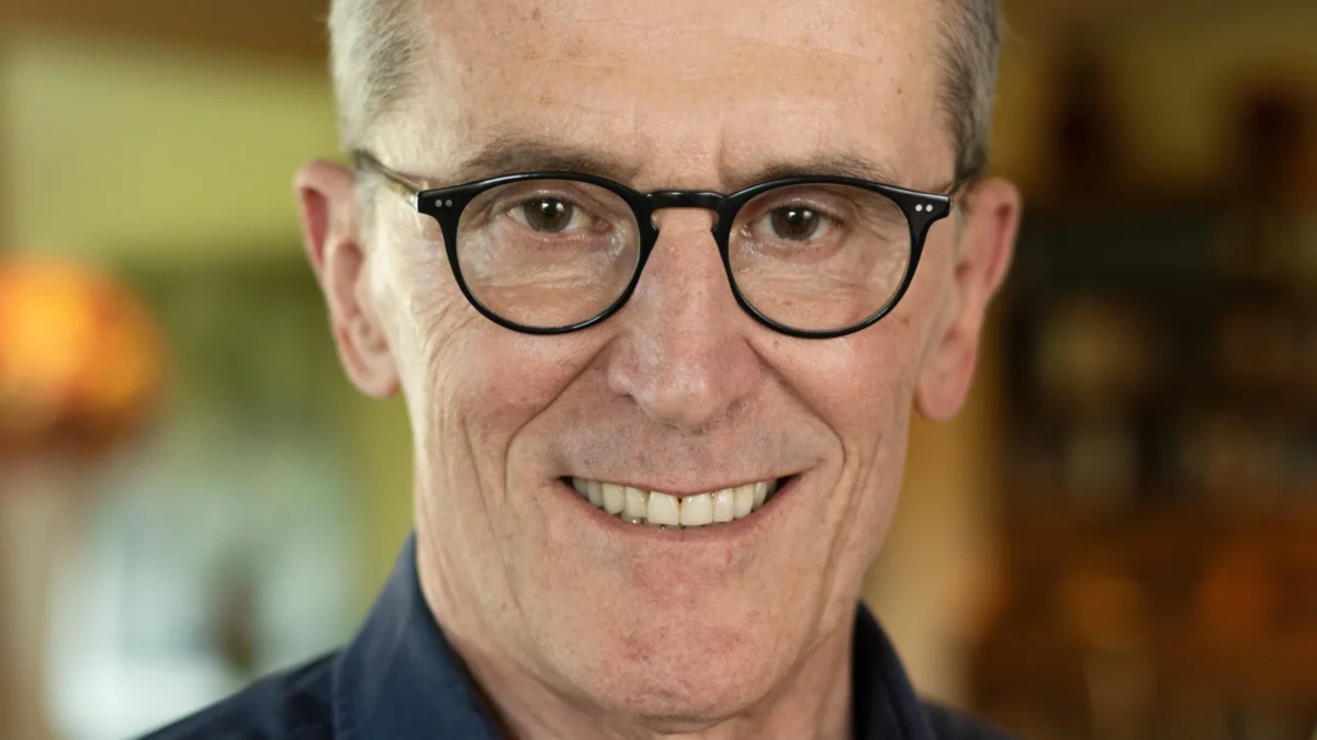 Close-up of a smiling older man wearing black glasses against a blurred indoor background.