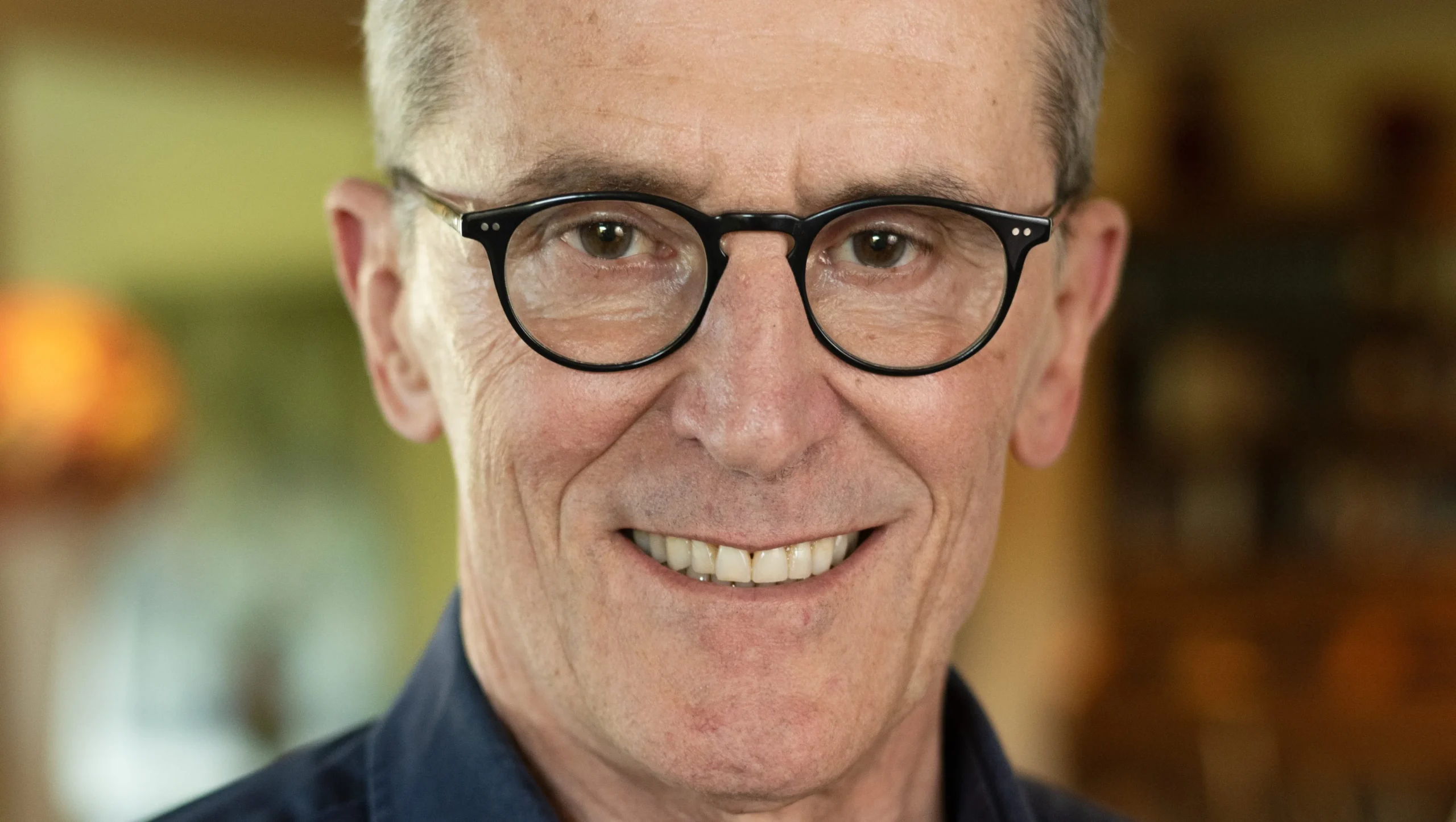 Close-up of a smiling older man wearing black glasses against a blurred indoor background.