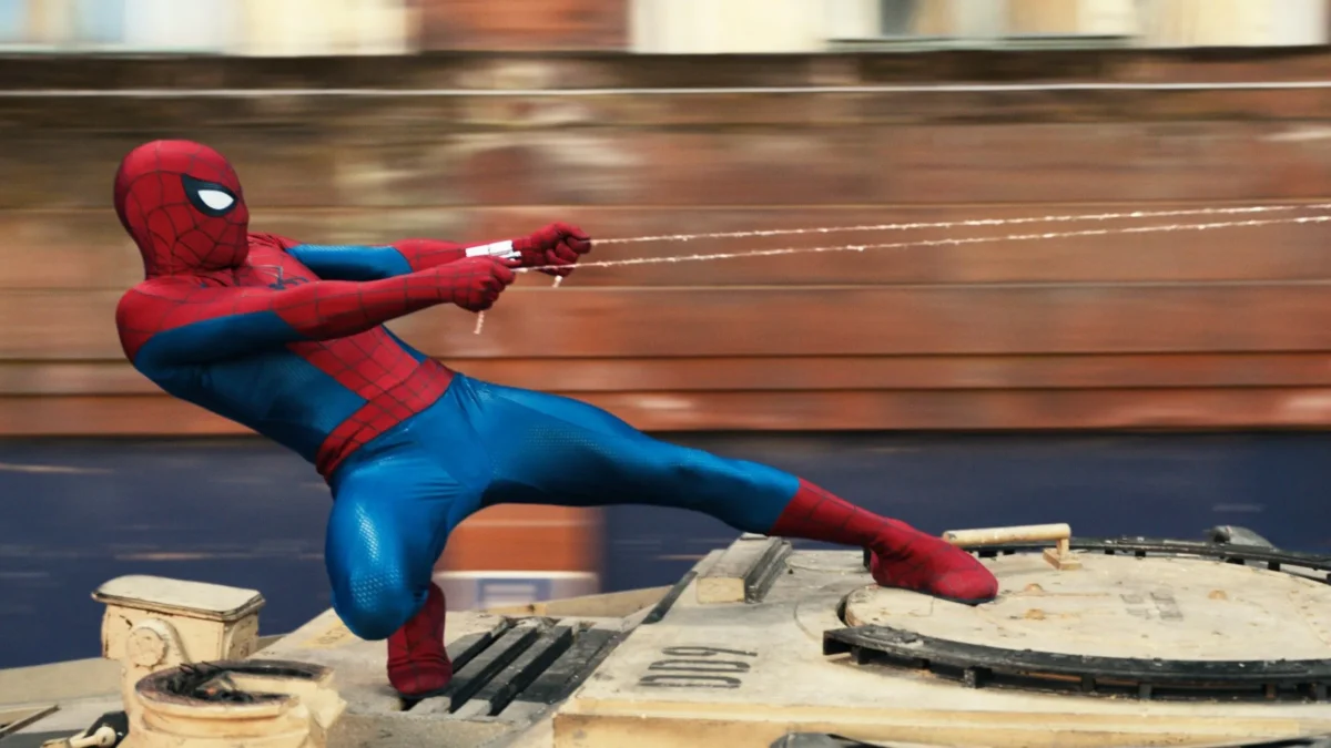 A masked superhero in a red and blue suit shoots webbing while crouched on a vehicle.