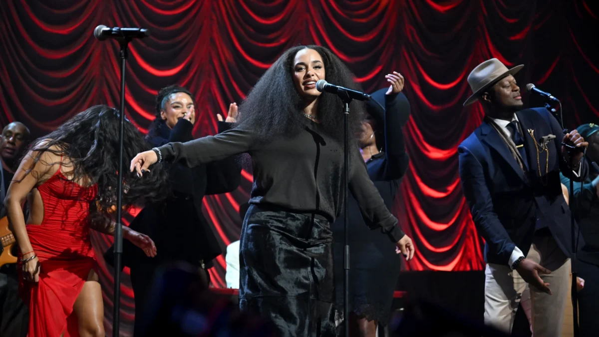 A singer performs at a microphone with dancers and musicians before red draped stage curtains.