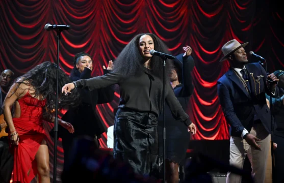 A singer performs at a microphone with dancers and musicians before red draped stage curtains.