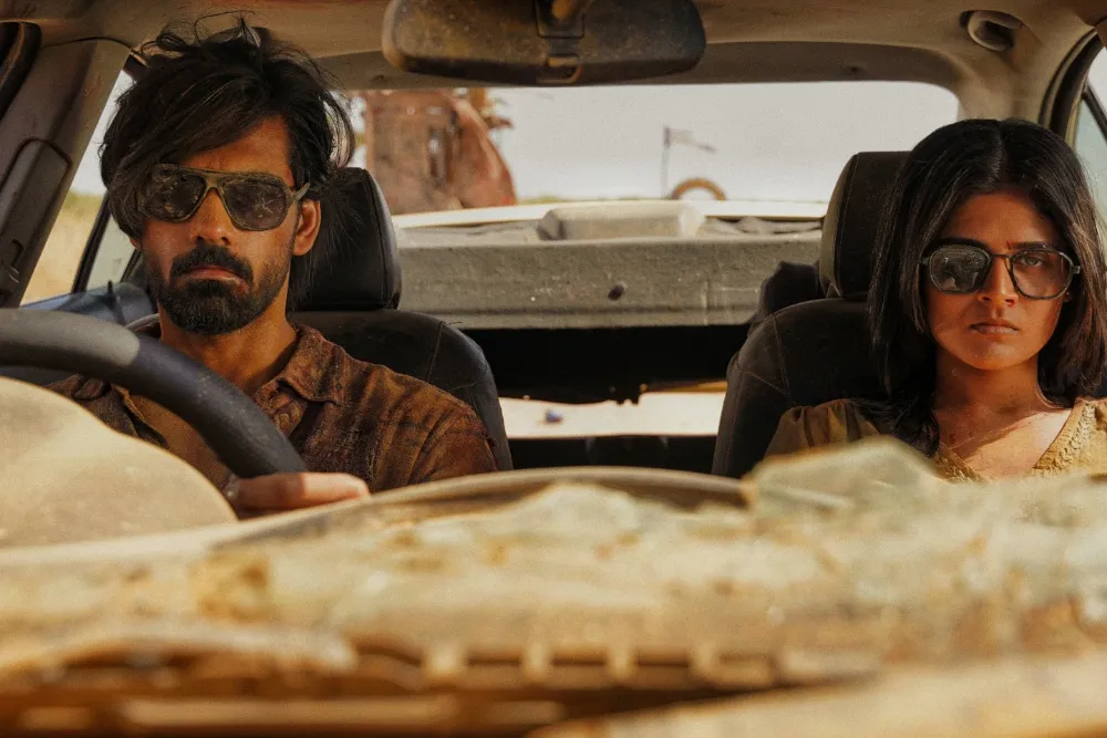 Two people wearing sunglasses sit in a dusty car, seen through the windshield.