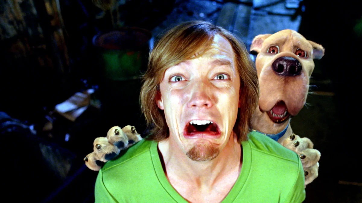 A frightened man in a green shirt grimaces as a large dog behind him opens its mouth.