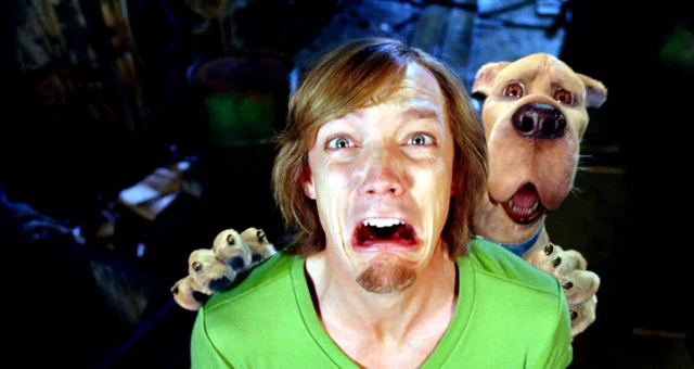 A frightened man in a green shirt grimaces as a large dog behind him opens its mouth.