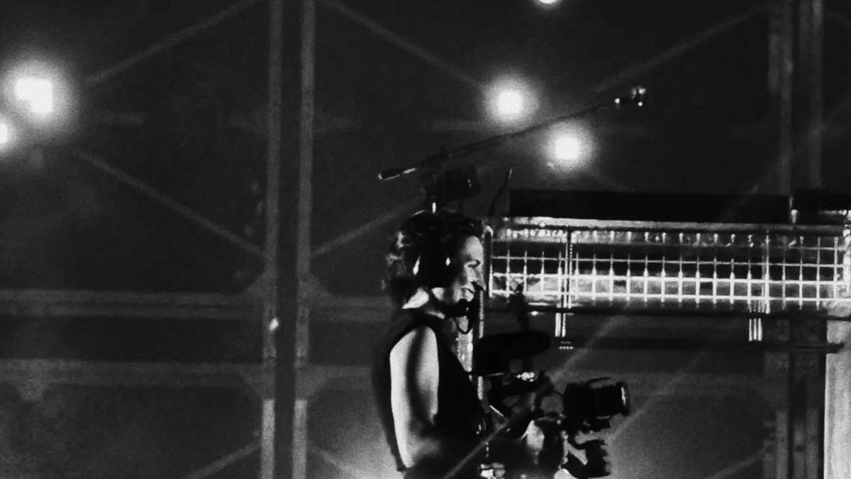A person wearing a headset carries a large camera rig amid bright lights and metal scaffolding.