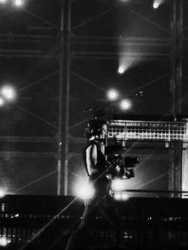A person wearing a headset carries a large camera rig amid bright lights and metal scaffolding.
