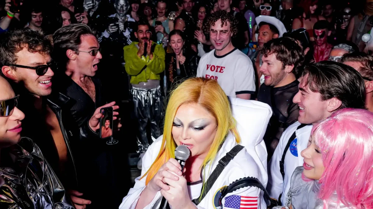 A performer in a white astronaut costume sings into a microphone amid a crowded, colorful party scene.