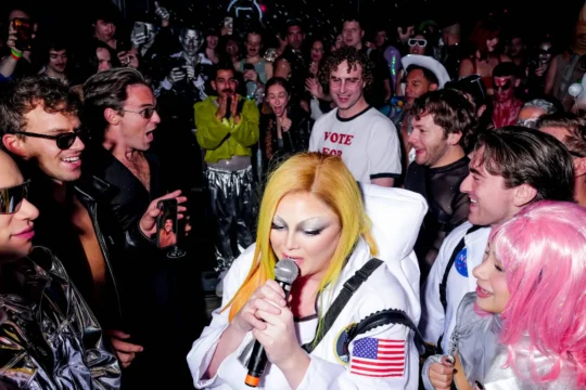 A performer in a white astronaut costume sings into a microphone amid a crowded, colorful party scene.