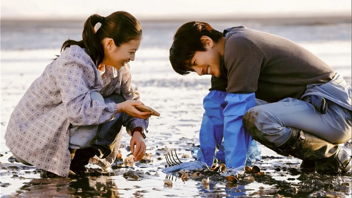 Two people crouch on a muddy shoreline, smiling while collecting shellfish with gloves and a rake.