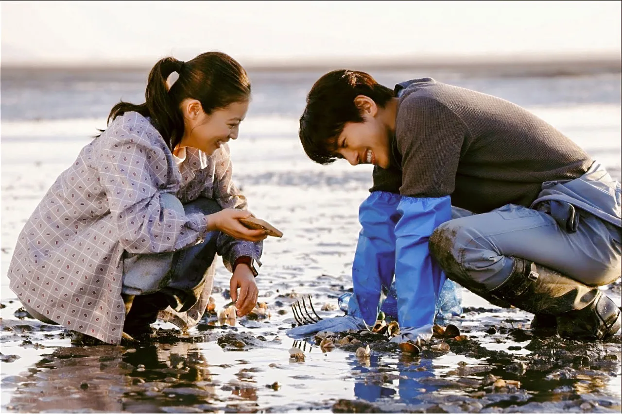 Two people crouch on a muddy shoreline, smiling while collecting shellfish with gloves and a rake.