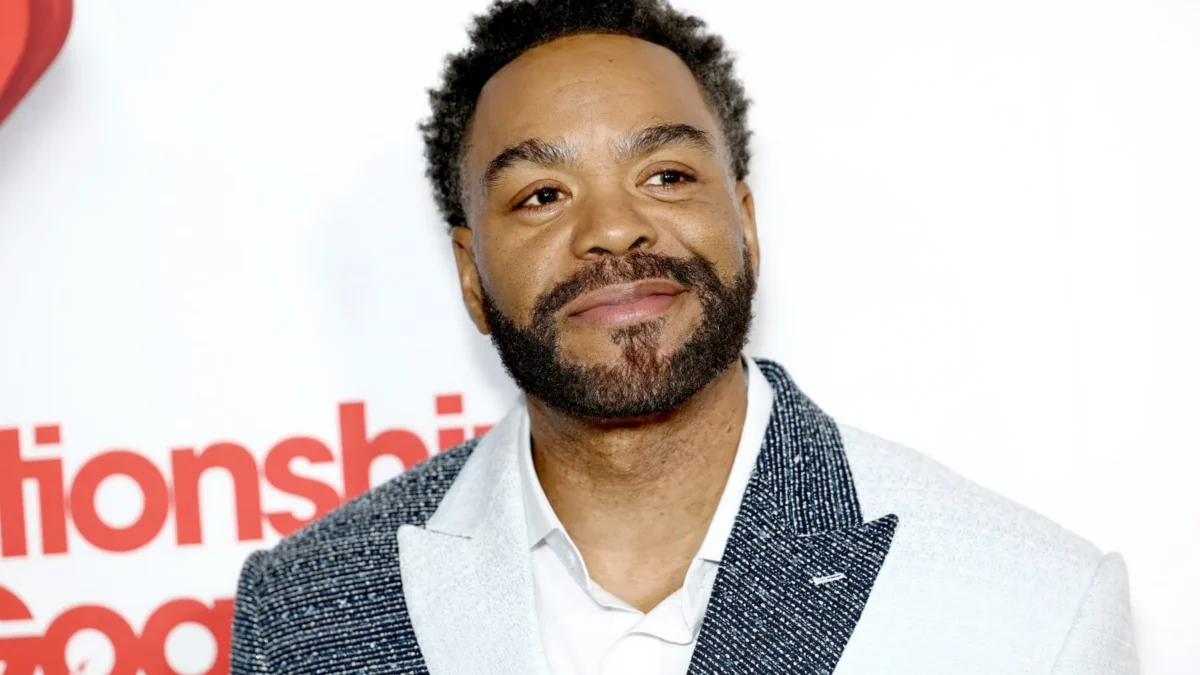 A smiling bearded man in a patterned blazer poses against a white backdrop with red lettering.