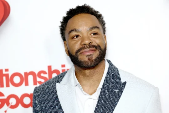 A smiling bearded man in a patterned blazer poses against a white backdrop with red lettering.