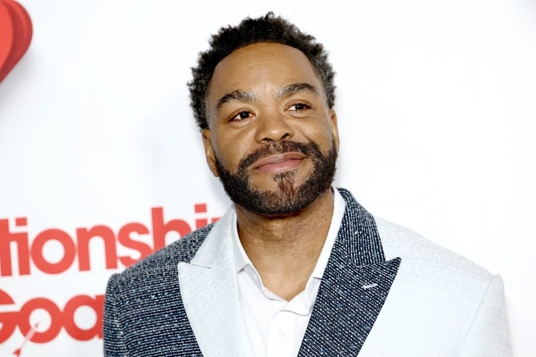 A smiling bearded man in a patterned blazer poses against a white backdrop with red lettering.