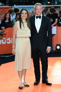Two people pose on an orange carpet, one in a pale dress and one in a black tuxedo.