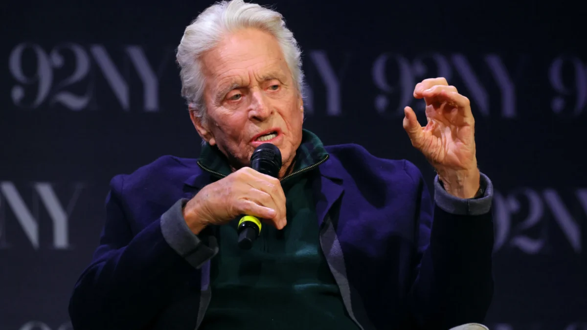 Older white-haired man seated with a microphone, raising one hand while speaking against a dark backdrop.