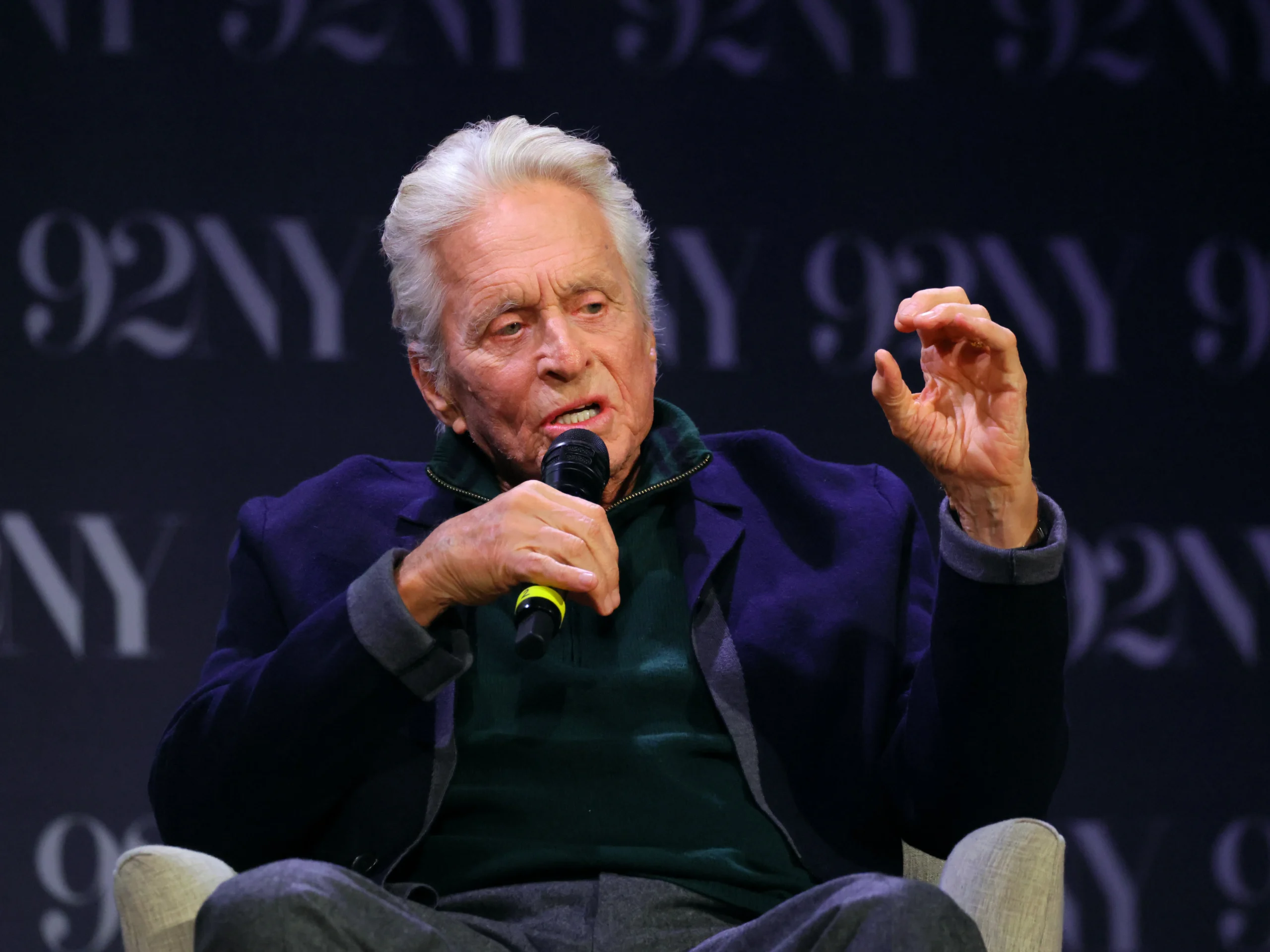 Older white-haired man seated with a microphone, raising one hand while speaking against a dark backdrop.