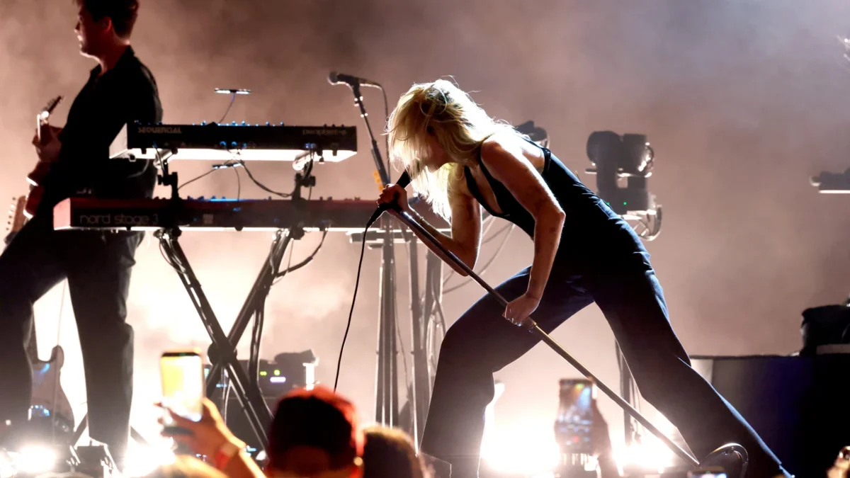 A blonde person leans into a microphone stand on a smoky, backlit stage beside another musician.