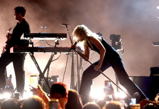 A blonde person leans into a microphone stand on a smoky, backlit stage beside another musician.