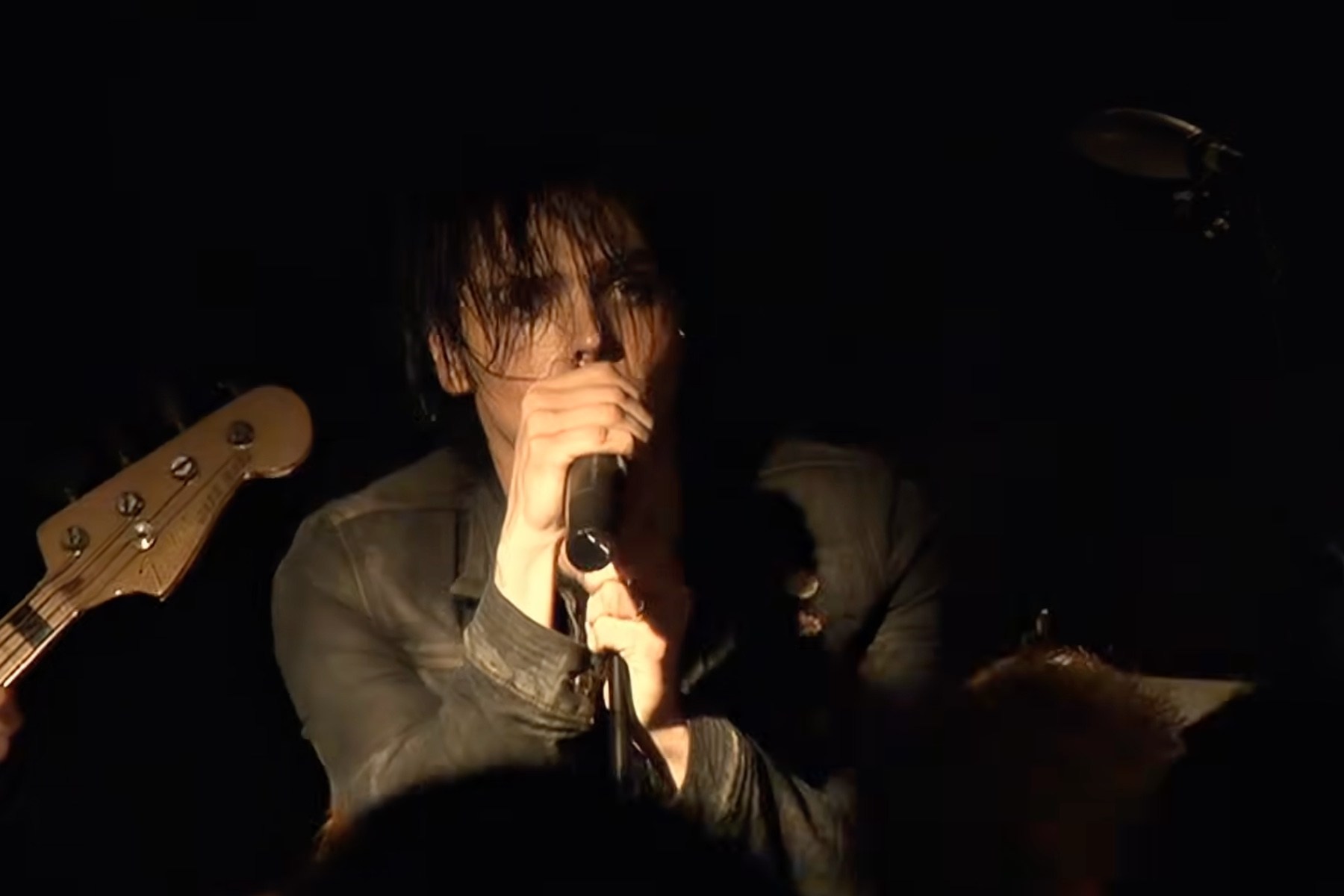 A dark-haired singer holds a microphone on a dimly lit stage, with a guitar neck visible beside them.