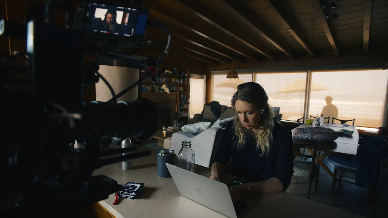 A camera films a woman working on a laptop in a dim room overlooking a beach.