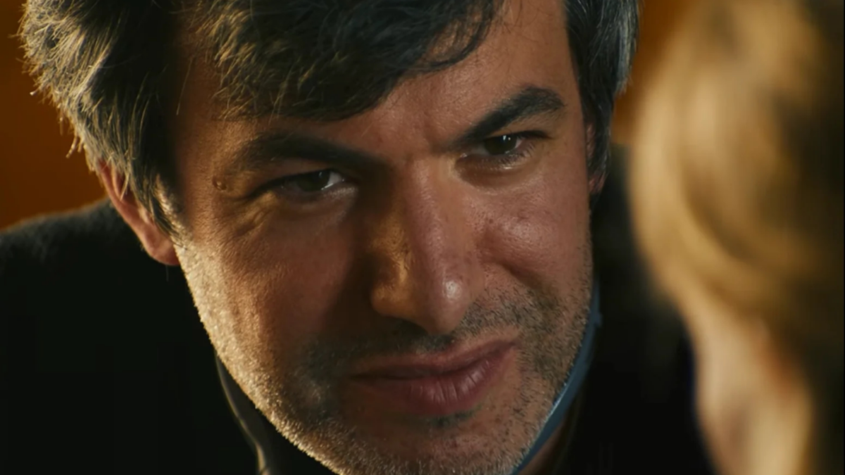 A close-up of a dark-haired man with stubble looking toward a blurred blonde person in warm light.