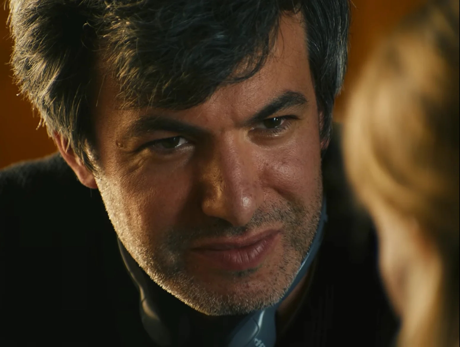 A close-up of a dark-haired man with stubble looking toward a blurred blonde person in warm light.