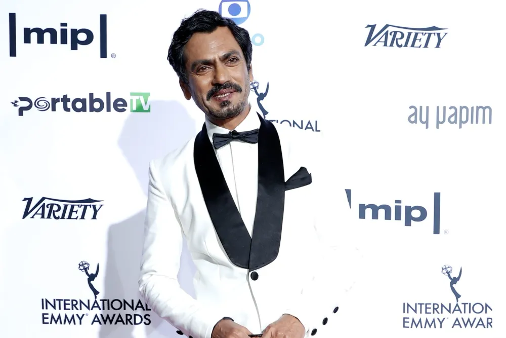 A man in a white tuxedo and black bow tie poses before a logo-covered awards backdrop.