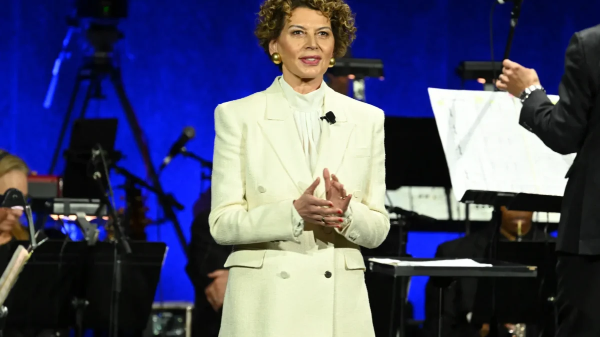 A woman in a white suit stands applauding on a blue-lit stage with musicians and microphones behind her.