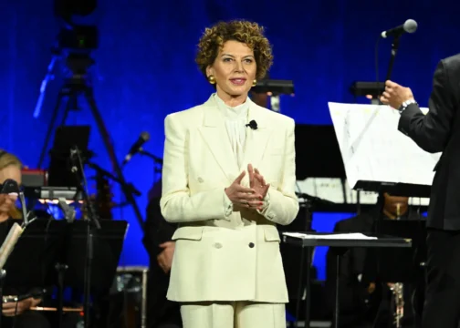 A woman in a white suit stands applauding on a blue-lit stage with musicians and microphones behind her.