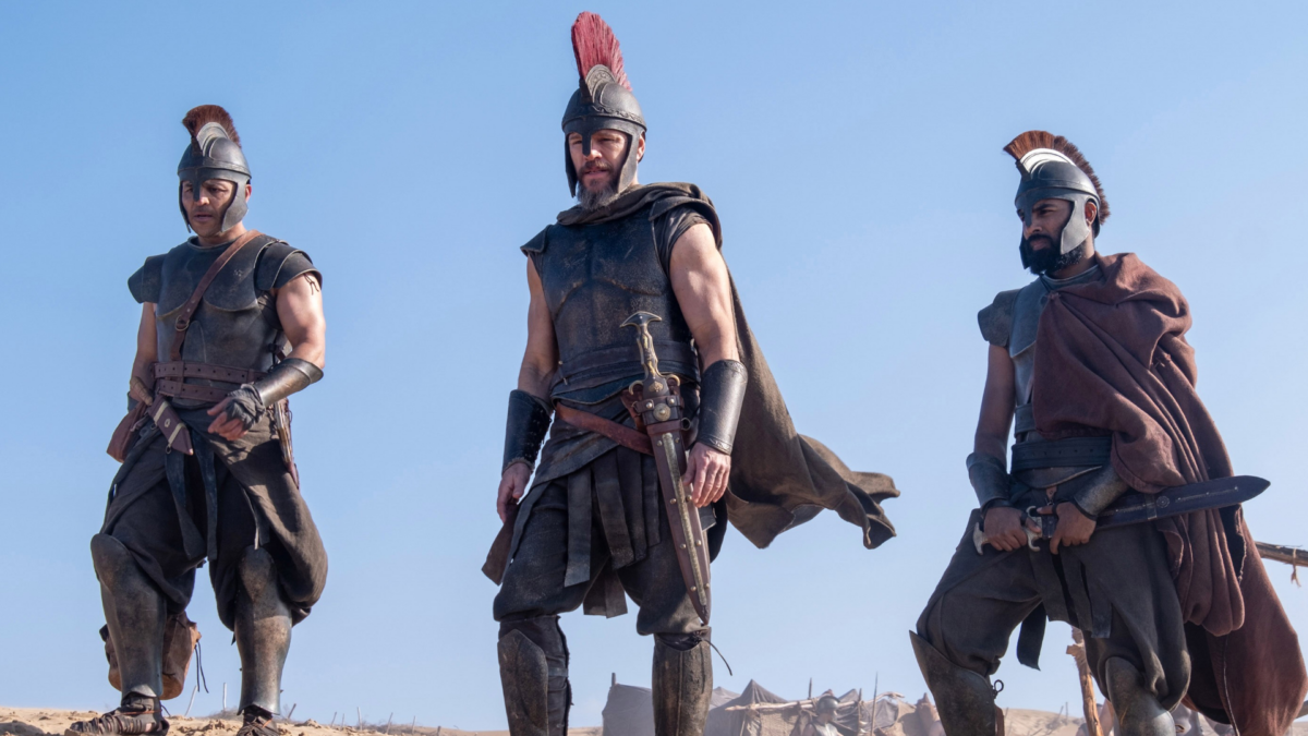 Three armored warriors in crested helmets stand on sandy ground under a clear blue sky.