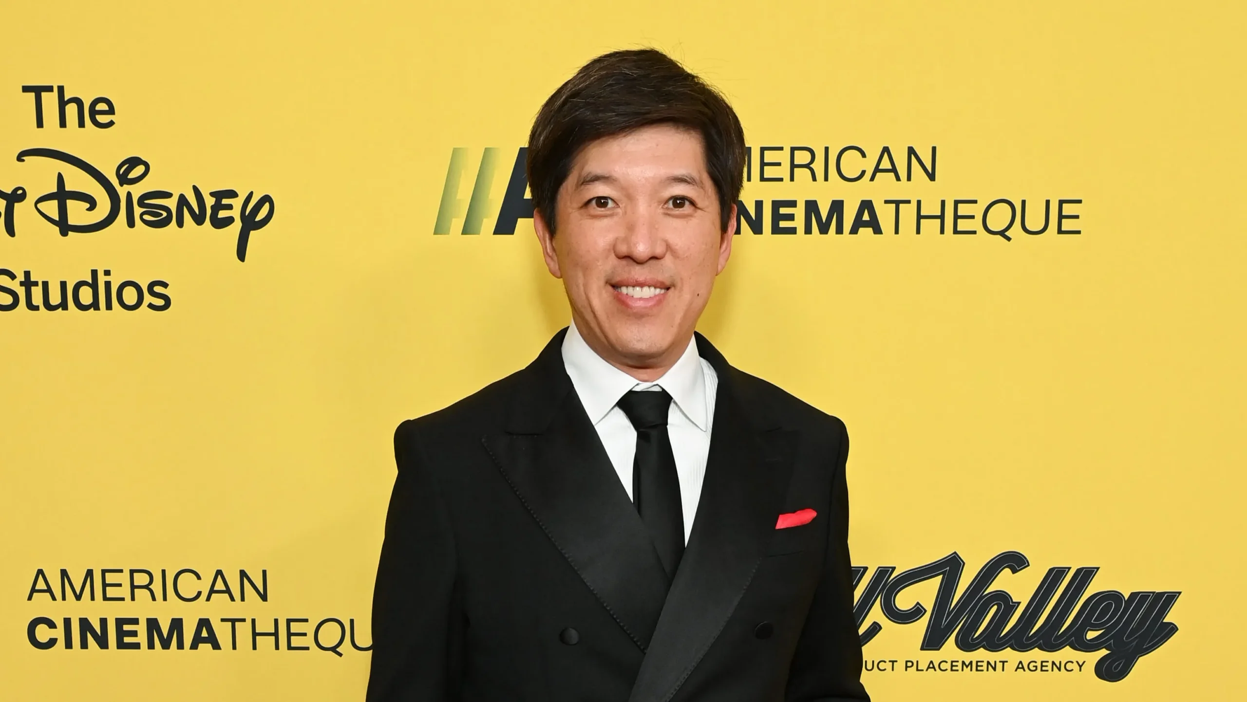 A man in a black suit and tie smiles in front of a yellow backdrop with event logos.