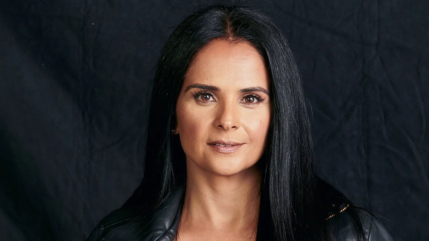 Woman with long black hair in a black jacket poses against a dark textured background.