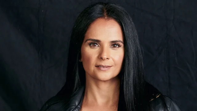 A woman with long black hair wearing a black jacket poses against a dark textured background.
