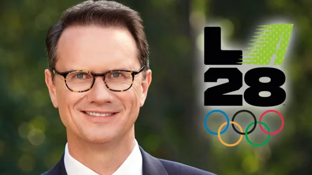 A smiling man in glasses and a suit appears beside the LA28 logo and Olympic rings.