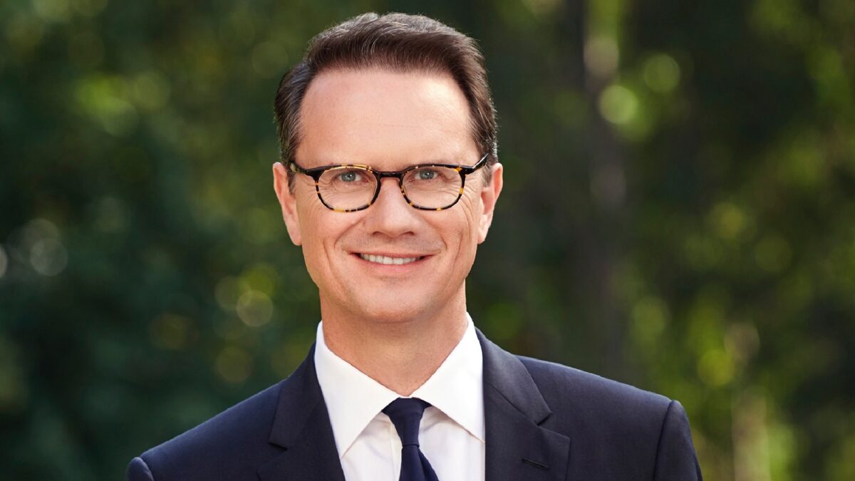 A smiling man in glasses and a dark suit stands before a blurred green background.