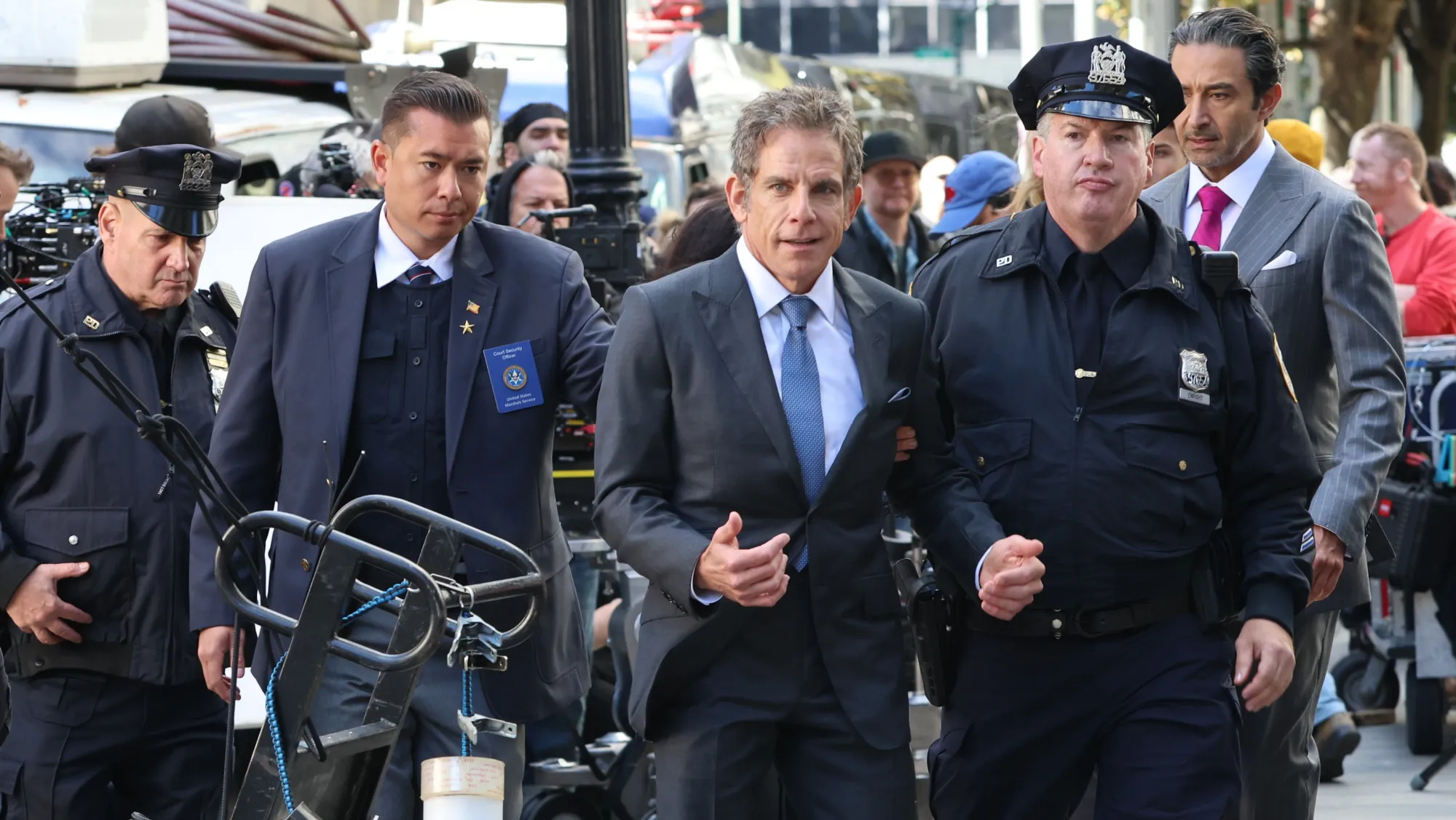 Uniformed officers and suited men walk together past cameras and equipment on a crowded city sidewalk.