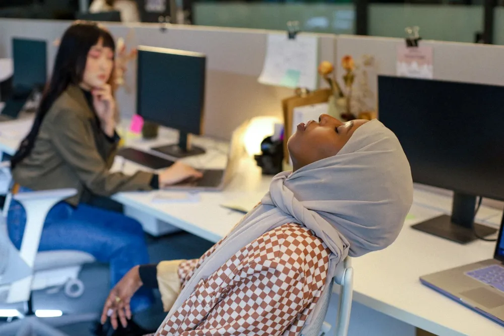 A person in a light headscarf leans back at an office desk while another person works nearby.