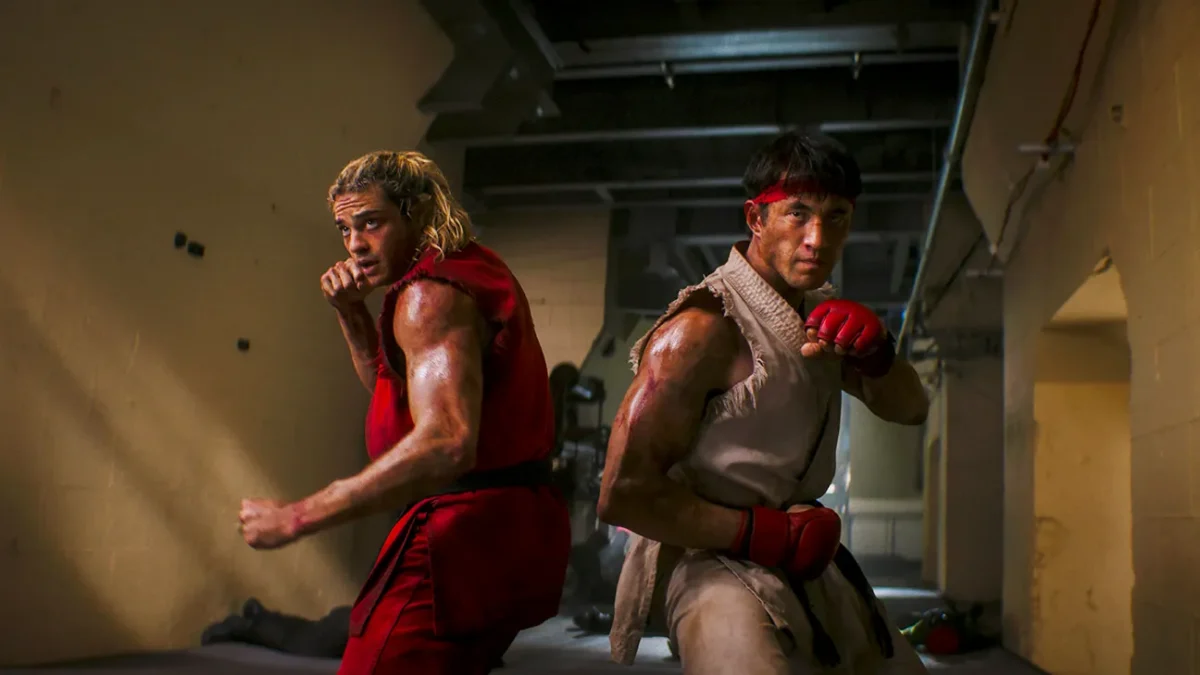 Two men in sleeveless martial arts outfits pose with raised fists in a dim, industrial hallway.