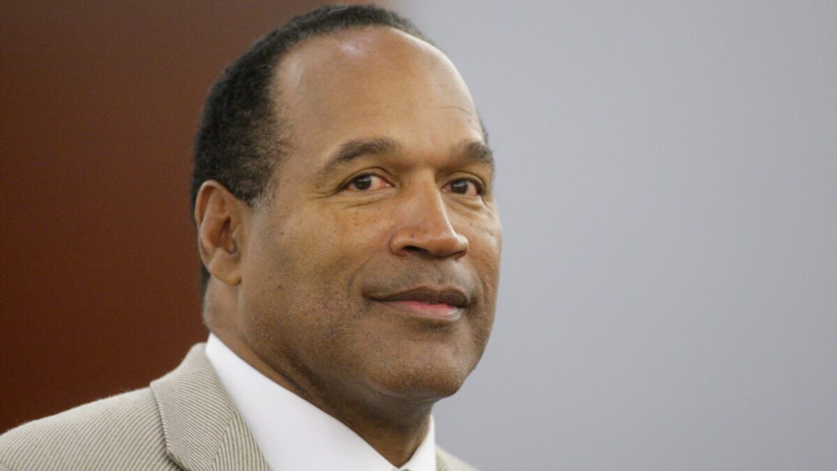 Close-up of a man in a beige suit and striped tie against a plain background.