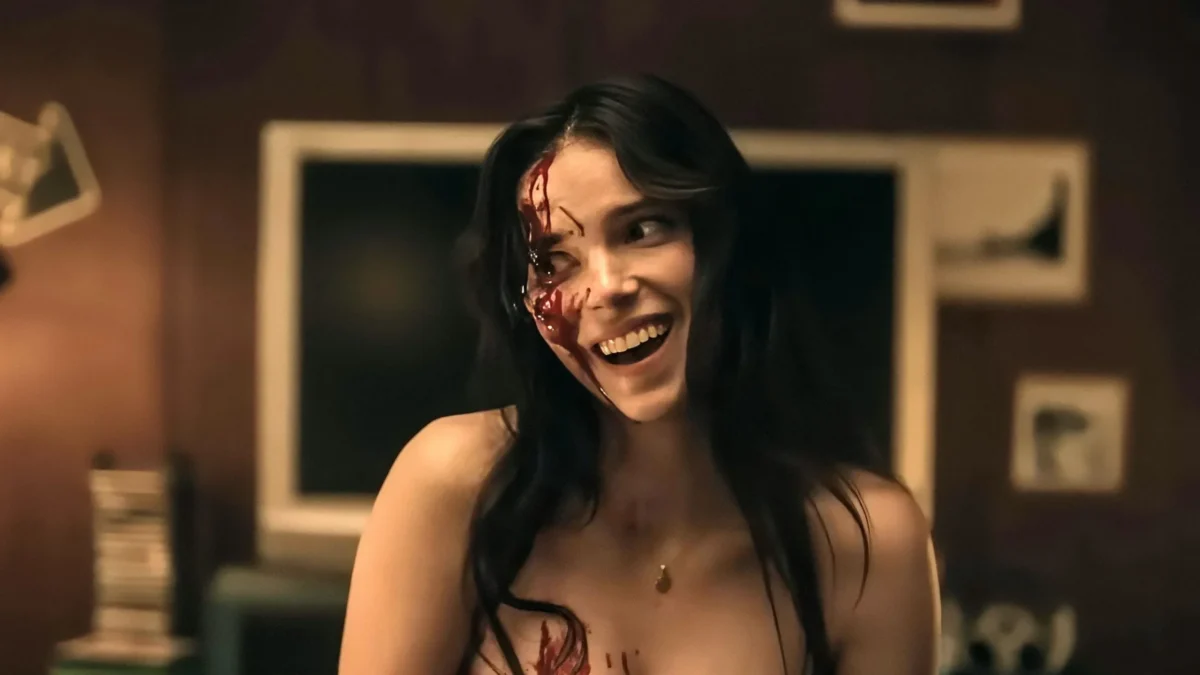 A smiling woman with long dark hair has red liquid on her face and chest in a dim room.