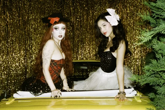 Two people in bow-accented outfits sit in a convertible before a glittering gold tinsel backdrop.