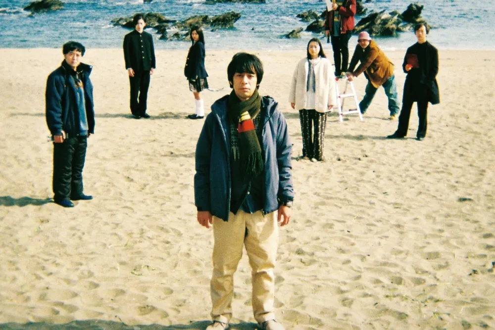 Several people stand spaced apart on a sandy beach, with rocks and ocean waves behind them.