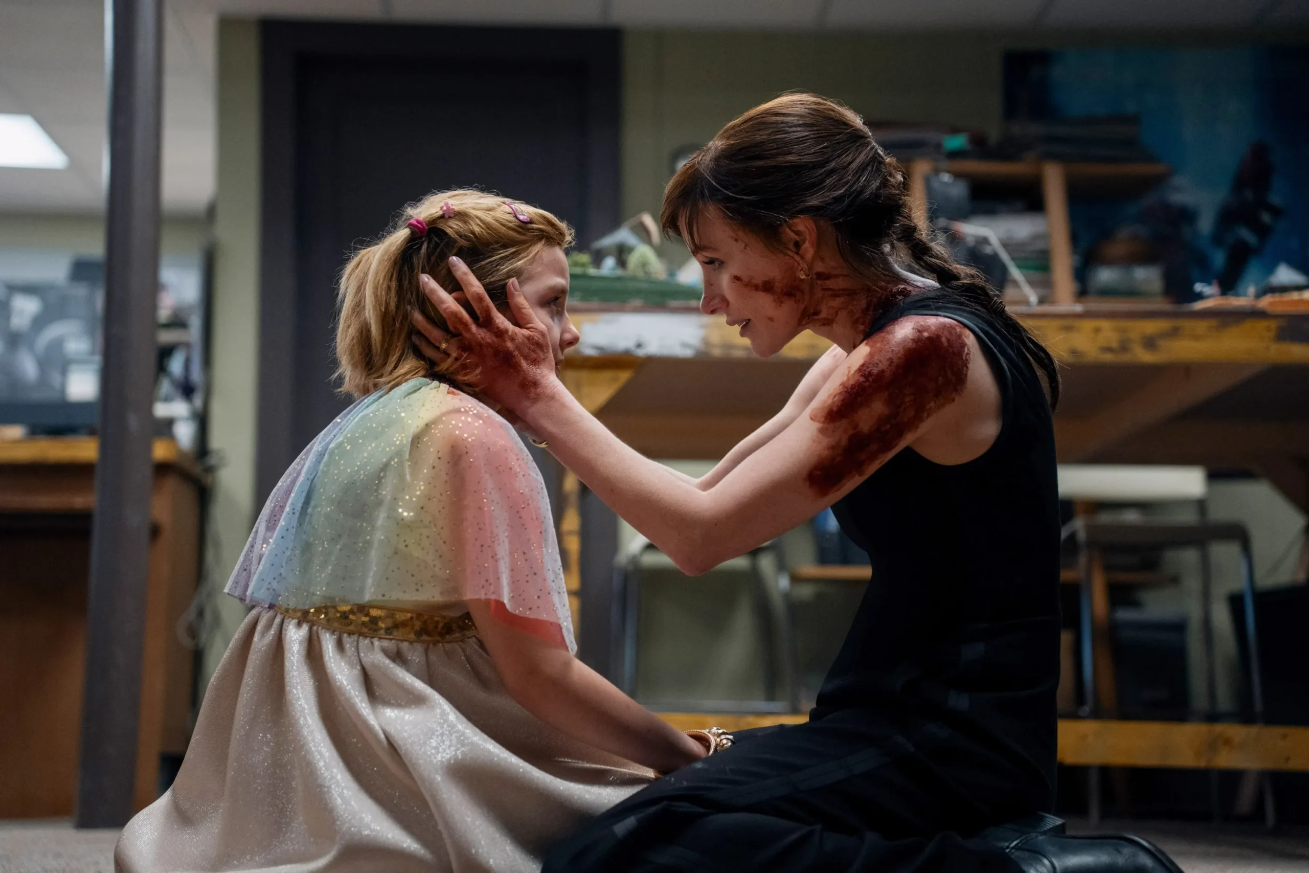 A blood-streaked woman kneels, holding a child's face in a dim room with desks.