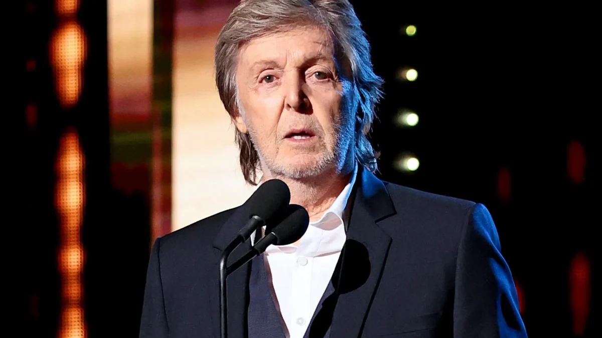 Older man in a dark suit speaks at a microphone on a stage with colorful lights.
